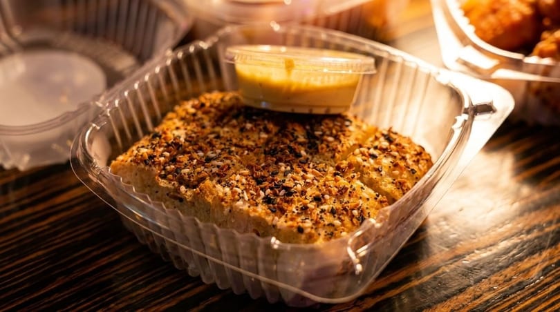 Everything bagel seasoned breadsticks with a side of dipping sauce in a plastic takeout container.