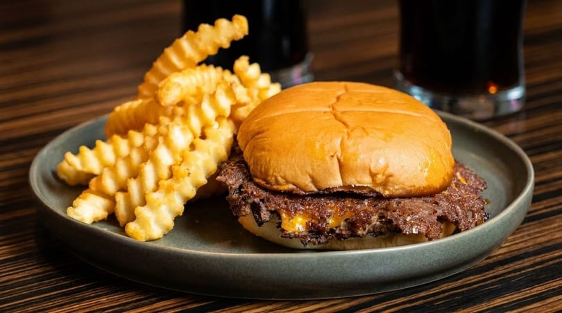 A smash burger with melted cheese and crinkle-cut fries on a gray plate, with drinks in the background.