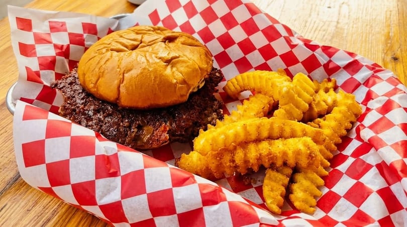 A burger with a thick patty and crinkle-cut fries in a red and white checkered paper-lined basket.