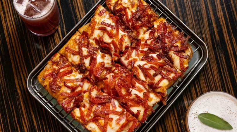 Square-cut pizza with melted cheese and crispy pepperoni strips, served with two drinks on a dark wooden table.